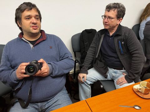 Dva sedící účastníci jednání, jeden drží fotoaparát